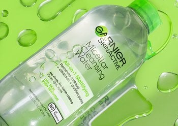 Top shot of Micellar Cleansing Water Mattifying, green cap, bottle. The bottle is lying horizontally on a green surface, on top of large water droplets.