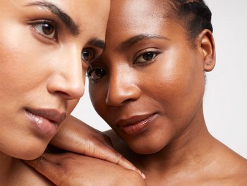 A close-up of two women with different skin tones, looking towards the camera with soft expressions, suggesting unity or a natural look.