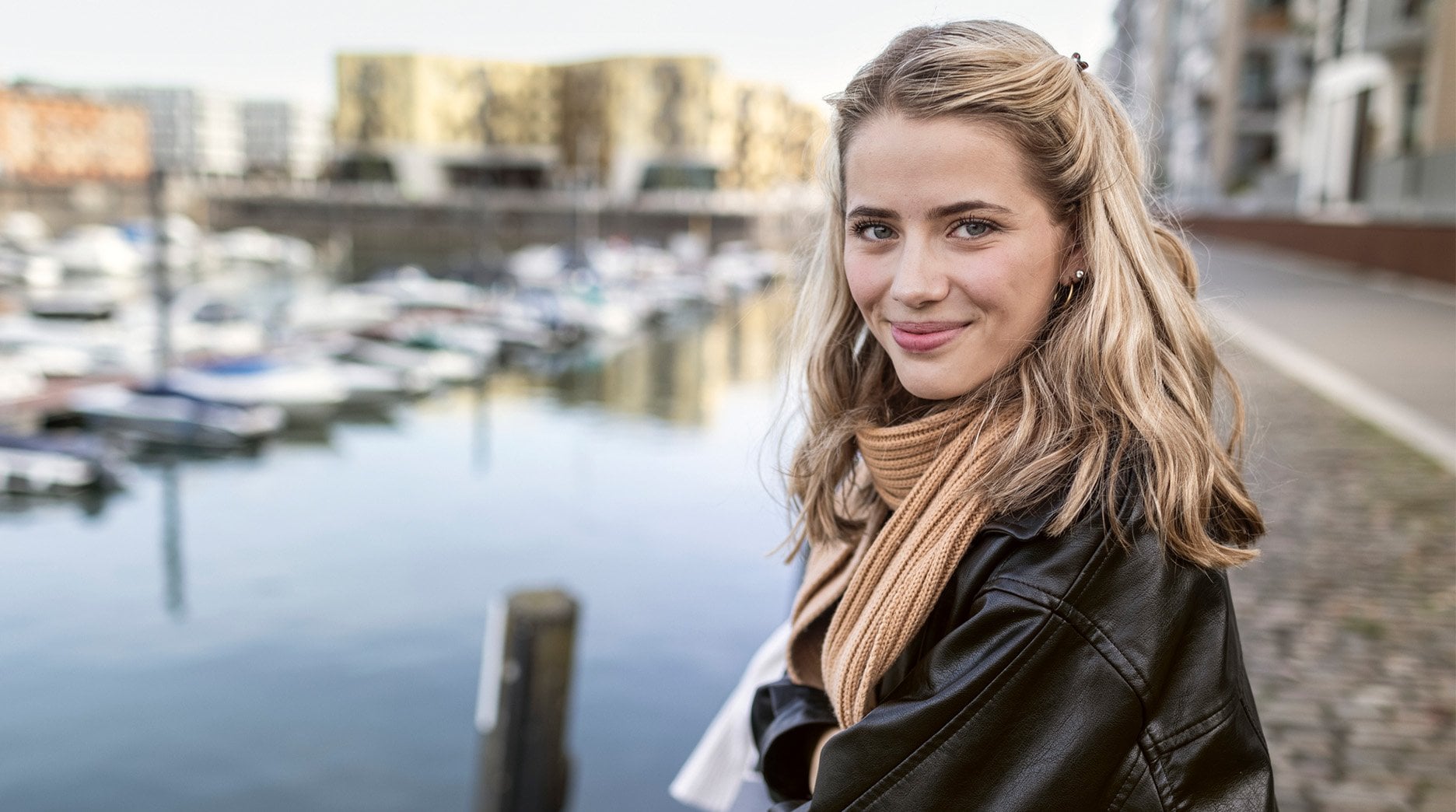 Portrait Of Smiling Young Woman Leaning On Railing At Urban Marina. She has healthy blond mid-length hair.