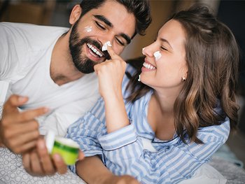 A happy couple in bed, playfully applying face cream to each other's noses, laughing and enjoying a skincare moment together.