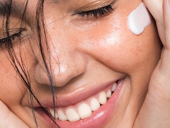 A close-up of a person with wet, dark hair and a joyful expression, applying a white cream to their face near their eye.