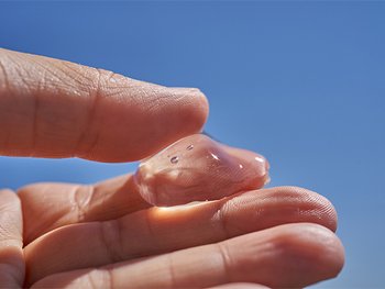 A close-up of a hand holding a translucent, jelly-like substance with small bubbles, against a clear blue sky background.