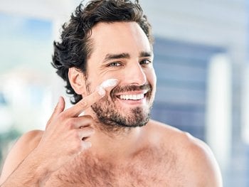 A smiling man applies moisturizer to his face, depicting the act of moisturizing.