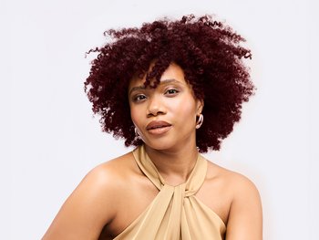A woman with voluminous, reddish-brown curly hair and warm makeup, wearing a gold halter-neck top, looking towards the camera against a white background.