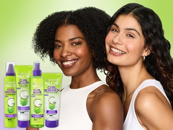 Two diverse females smiling at the camera. One has coily hair and the other wavy long hair. Line up of Curl Nourish products is featured to the left side of the photo.