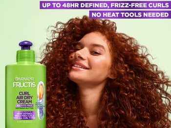 A Garnier Fructis Curl Air Dry Cream bottle alongside a woman with voluminous curly hair. The image includes text claiming "UP TO 48HR DEFINED, FRIZZ-FREE CURLS" and "NO HEAT TOOLS NEEDED".