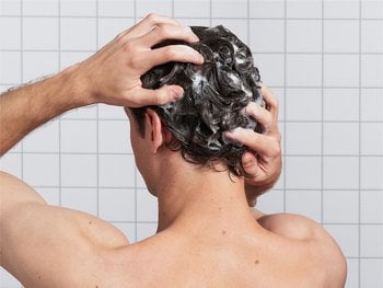 A person with short brown hair is shown from the back, lathering their hair with shampoo.