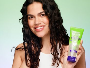 Brunette model smiling at camera holding a bottle of Garnier Fructis Scrunch Controlling Gel.