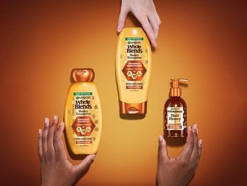 Top view photo of Honey Treasure Repairing System products laid out on a brown/yellow honey-colored background, with hands of different ethnicities reaching out grabbing each product. Honey Treasure Repairing Shampoo, Repairing Conditioner, and Repairing Serum are featured.