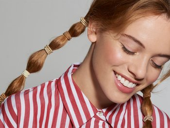 A woman with long, light brown hair smiles as she showcases a bubble ponytail adorned with small, gold elastics.