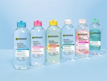 A row of six different Garnier Micellar Cleansing Water bottles, each with a different colored cap (blue, pink, light green, light blue, peach, light green again) and variations in formula, on a light blue surface.